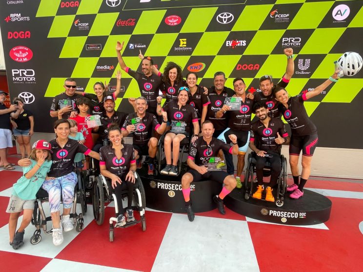 Foto de un grupo de personas con y sin discapacidad, en el pódium del circuito de Montmelo con los maillots de DiscaSport, al final de la carrera del Gran Premi Isidre Esteve 2023.